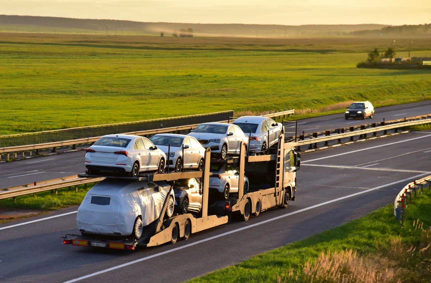 A wide shot of a car carrier trailer truck transporting cars on a highway against an open field and a sunset background.