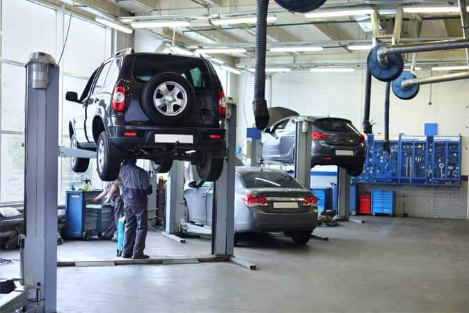 A wide shot of an auto repair shop garage with two mechanics working on three cars.